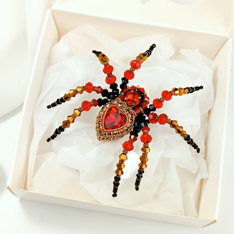 May include: A red and black beaded spider brooch with a large red heart-shaped rhinestone in the centre. The spider is set in a white box with white tissue paper.