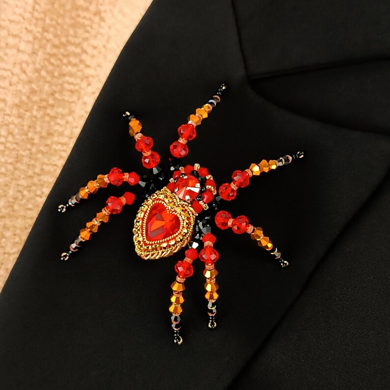 May include: A red and black beaded spider brooch with a large red heart-shaped gem in the centre. The spider is made of small beads and has a detailed design.