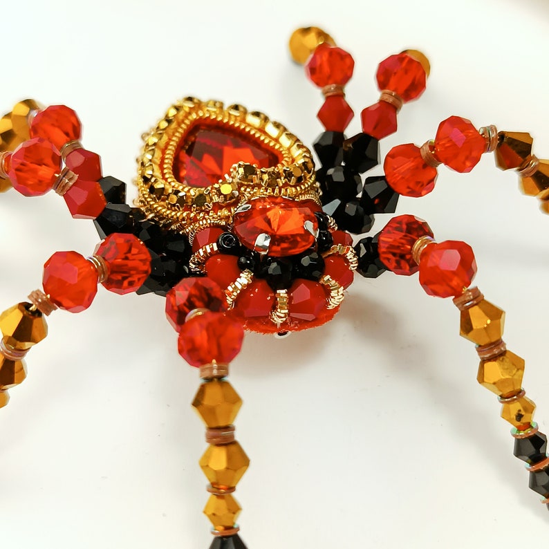 May include: A beaded spider brooch featuring a red, black and gold colour scheme. The spider's body is decorated with a large red jewel and gold accents.