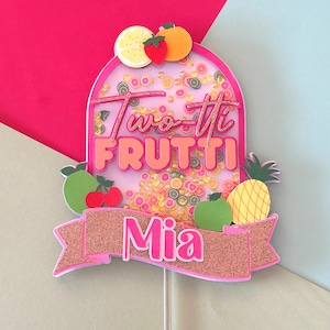 May include: A vibrant cake topper with the words "Two-tti Frutti" in pink and gold glitter. It includes a banner with the name "Mia" and is adorned with fruit illustrations, such as a lemon, strawberry, and orange.