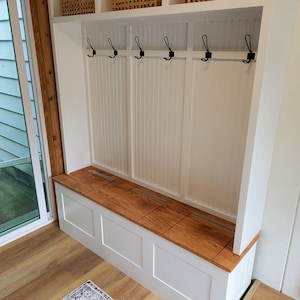 May include: White entryway bench with storage compartments and hooks. The bench has a wooden top and is built into a white cabinet with two shelves. The shelves are empty except for two woven baskets.