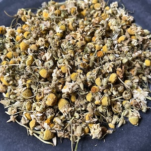 May include: Dried chamomile flowers in a gray bowl. The flowers are a light brown color with yellow centers.