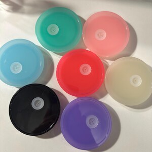 May include: A collection of seven colorful, round plastic lids. The lids are in various colors, including blue, green, pink, red, yellow, black, and purple. Each lid has a white, cross-shaped center. The lids are arranged in a circular pattern.