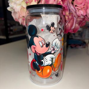 May include: A clear glass tumbler with a black lid, featuring a colorful Mickey Mouse design. The cartoon character is depicted holding a flower, with red stars and Mickey Mouse silhouettes scattered around the glass. The tumbler is filled with white tissue paper.