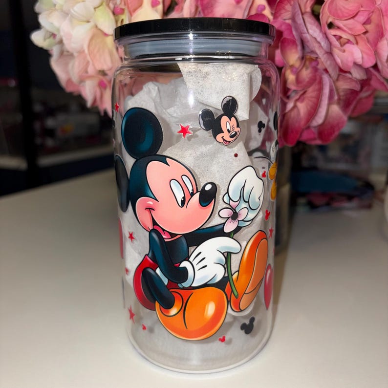 May include: A clear glass tumbler with a black lid, featuring a colorful Mickey Mouse design. The cartoon character is depicted holding a flower, with red stars and Mickey Mouse silhouettes scattered around the glass. The tumbler is filled with white tissue paper.