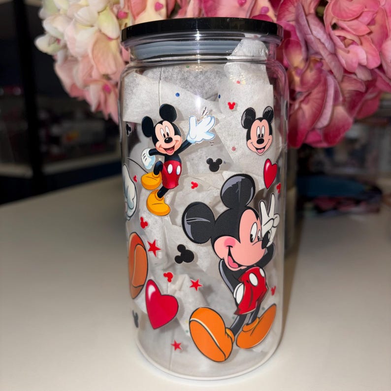 May include: A clear glass jar with a black lid, adorned with vibrant Mickey Mouse illustrations, hearts, and stars. The jar is filled with white tissue paper and is placed on a white surface.