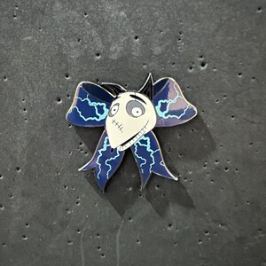 May include: A blue and white enamel pin featuring a character from the movie "The Nightmare Before Christmas". The character is wearing a blue bow with lightning bolt designs.