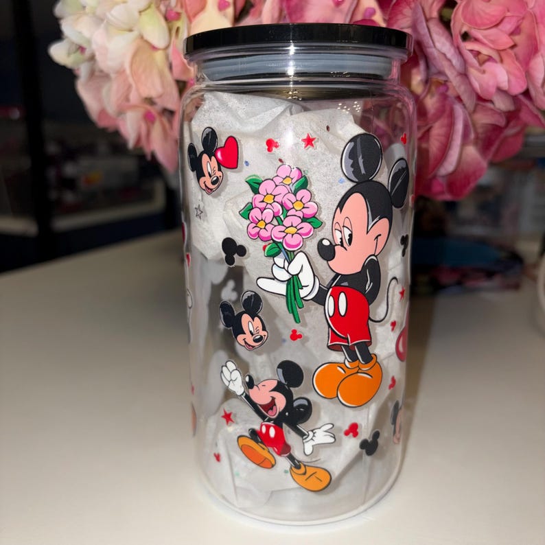 May include: A clear glass jar with a black lid, decorated with colorful images of Mickey Mouse. Mickey is holding a bouquet of pink flowers. The jar is filled with white tissue paper and surrounded by pink flowers.