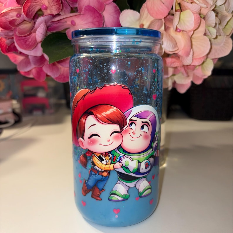 May include: A clear, cylindrical tumbler with a blue lid and base, featuring a cartoon illustration of two characters. The characters are hugging, with one wearing a cowboy hat and the other in a space suit. The tumbler is filled with blue glitter.