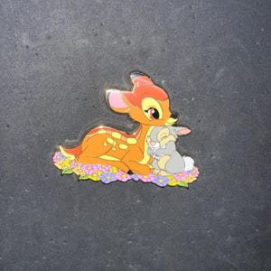 May include: A gold-tone pin featuring Bambi the deer from Disney's Bambi, hugging Thumper the rabbit. Bambi is orange and brown with white spots, and Thumper is gray. They are surrounded by pink and purple flowers.