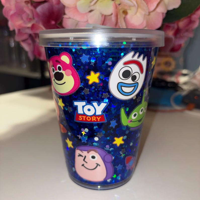 Może przedstawiać: Przezroczysty plastikowy kubek z niebieskim brokatowym tłem i wizerunkami postaci z Toy Story. Kubek posiada logo Toy Story oraz wizerunki Lotso, Forky'ego, Buzz'a Astrala i kosmity. Kubek posiada przezroczystą pokrywę.