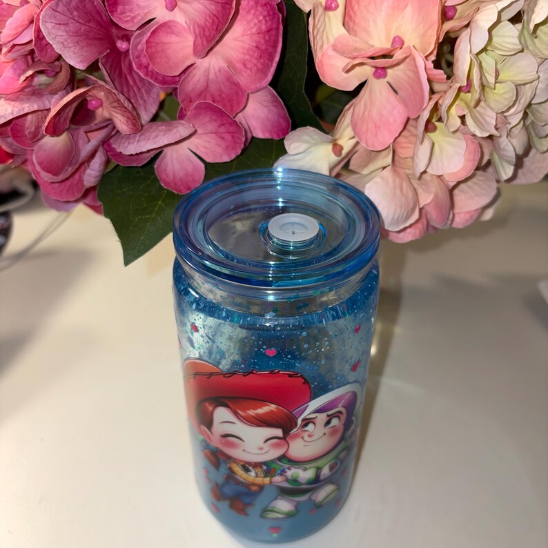 May include: A blue, glitter-filled tumbler featuring a cartoon image of Woody and Jessie from Toy Story. The tumbler has a blue lid and a clear body. Pink and white flowers are in the background.