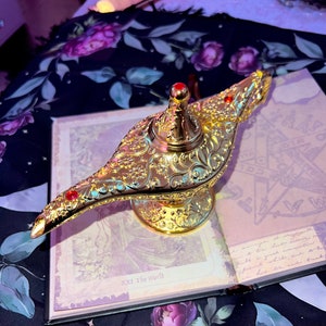 Puede incluir: Lámpara de genio dorada ornamentada con intrincados diseños florales y detalles de joyas rojas. La lámpara se encuentra sobre un libro abierto con imágenes de cartas del tarot. El fondo presenta una tela estampada floral.