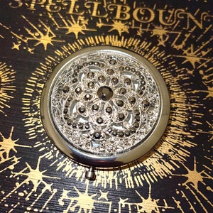 May include: A round, silver compact mirror with an ornate, openwork design. The mirror's surface is embellished with small, dark-colored stones. The background is black with gold starburst and text that reads "SPELLBOUND".