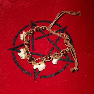 Puede incluir: Una pulsera hecha a mano con abalorios con forma de hueso, cuentas de madera marrón oscuro y un cordón tejido. La pulsera se muestra sobre una superficie roja con un diseño de pentagrama negro.