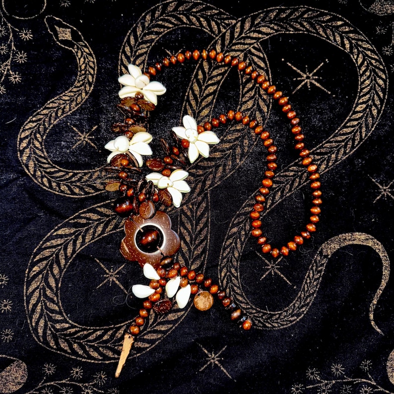May include: A handcrafted necklace featuring a combination of natural materials. The necklace incorporates wooden beads, white flower-shaped accents, and a central pendant with a flower design. The necklace is displayed on a black surface with a gold snake pattern.