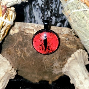 Puede incluir: Un collar con un colgante de ojo rojo en una cadena negra. El ojo está engastado en un cabujón de vidrio y tiene una pupila negra y un iris blanco.