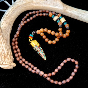 May include: A wooden bead necklace with a pendant. The pendant is gold-coloured with a horn-shaped design, featuring red, turquoise, and white enamel details. The necklace has a dark brown colour and is displayed on a black surface.