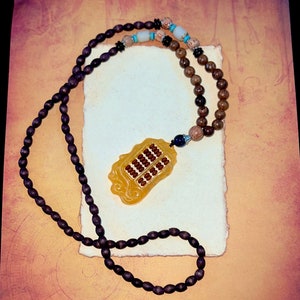 Può includere: Una collana di perline di legno marrone con un ciondolo a forma di abaco giallo. L'abaco è fatto di giada e ha perline marroni.