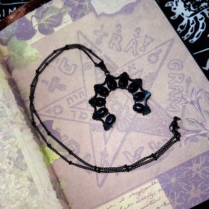 May include: A black necklace with a pendant featuring a horseshoe shape with oval-shaped black stones. The necklace is on a textured, light brown surface with black and white markings.