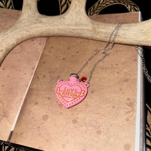 May include: A pink heart-shaped pendant necklace with the words "Love Potion" in gold lettering. The pendant has a silver chain and a small red bead. The necklace rests on a brown surface.