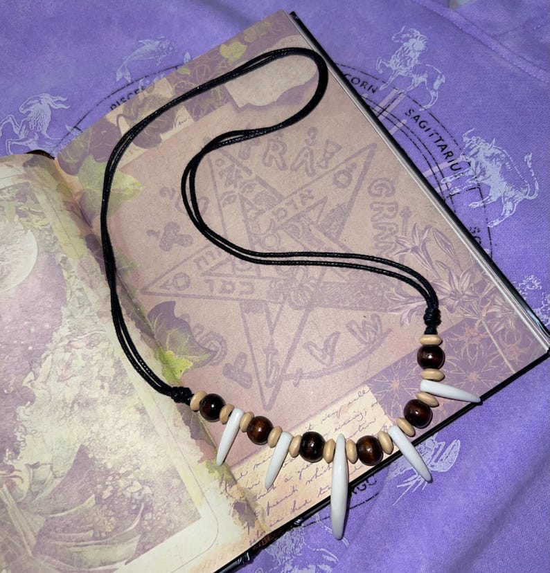 May include: A black cord necklace featuring a series of wooden beads, bone-colored spikes, and dark brown beads. The necklace is displayed on an open book with astrological symbols and text.