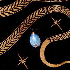 Può includere: Una collana con pendente a goccia blu opalescente, avvolta in filo di rame a forma di albero. Il pendente è appeso a un cordino nero su sfondo nero con motivi dorati e disegni a stella.