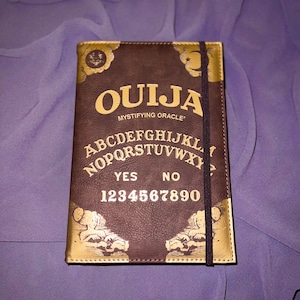 Puede incluir: Un diario encuadernado en cuero marrón con un diseño de tablero de Ouija de color dorado. El diseño incluye las letras del alfabeto, los números del 1 al 9 y el 0, las palabras "YES" y "NO", y las palabras "Mystifying Oracle".