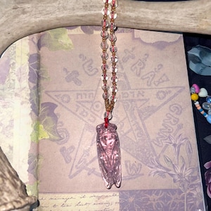 May include: A pink cicada pendant necklace with a beaded chain. The pendant is suspended from a red cord and hangs over an open book with text and a pentagram design. The background includes a deer antler and floral patterns.