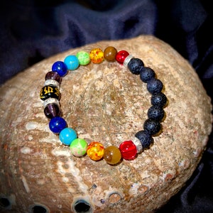 May include: A rainbow chakra bracelet with black lava rock beads and silver spacers. The bracelet features a variety of colorful gemstones, including turquoise, green, yellow, orange, red, and purple.