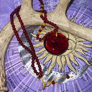 May include: A red glass pendant necklace with a beaded chain. The necklace is on a purple fabric background with a yellow and silver sun design.