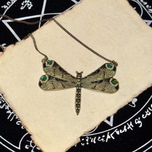 May include: A dragonfly pendant necklace featuring an antique bronze-toned metal frame with intricate filigree wing patterns, inlaid with dark green enamel, small white faux pearls, and sparkling green crystal accents on a thin metal chain.