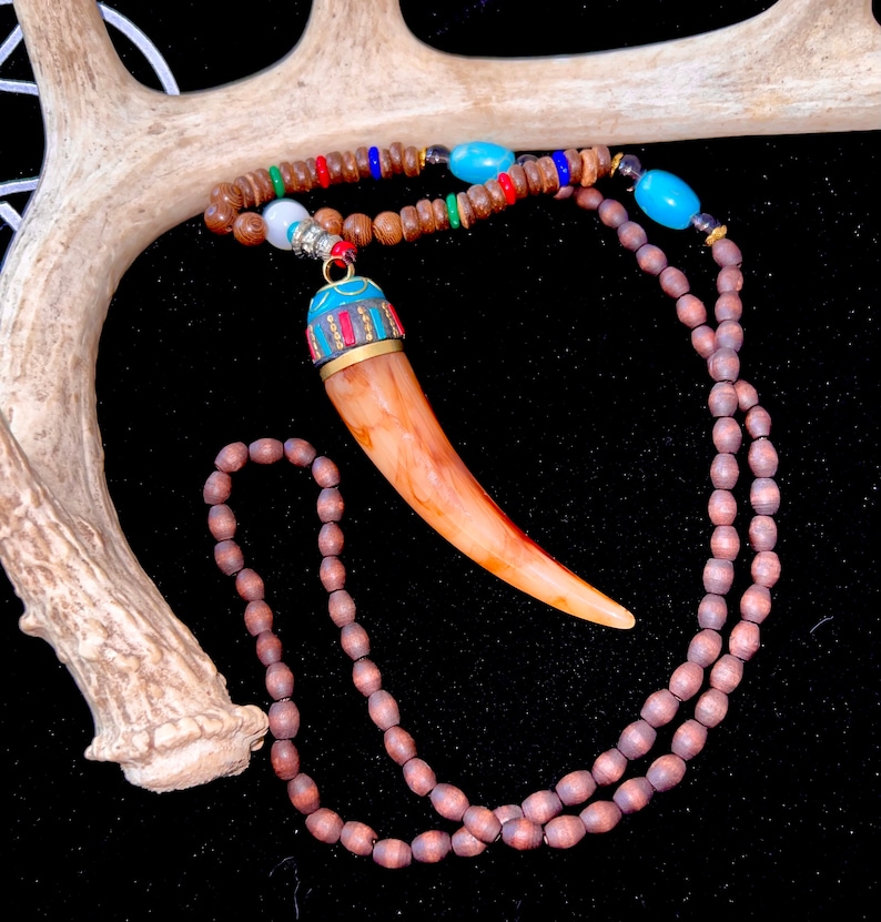 Op de afbeelding: Een bruine houten kralenketting met een bruine en blauwe gesneden hoorn hanger. De ketting is gedrapeerd over een wit hertengewei.