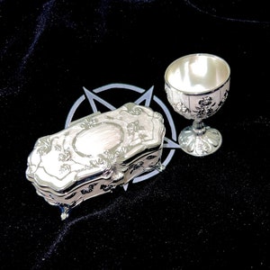 May include: A silver ornate box with a floral design and a silver chalice with a floral design on a black velvet background.