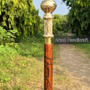 Op de afbeelding: Een houten wandelstok met een messing handvat. Het handvat heeft ingewikkelde bloemmotieven en een ronde bovenkant. De stok is bruin met een donker houtnerf.
