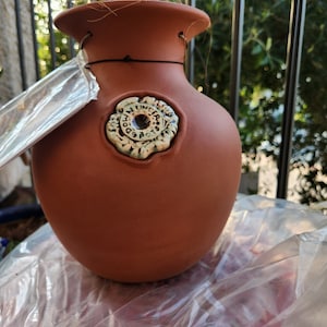 May include: A terracotta hanging planter with a decorative floral design and the words "The Flower Pot" in a circle. The planter is hanging from a black wire.