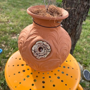 May include: A terracotta-colored, decorative birdhouse with a round body and a wide opening. The birdhouse features embossed floral patterns and a circular, decorative emblem with the text "Down Under Pots". A metal wire is attached for hanging.