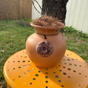 May include: A terracotta pot with a brown rope handle and a decorative design on the front. The pot is filled with brown coconut coir and is sitting on a yellow stool.