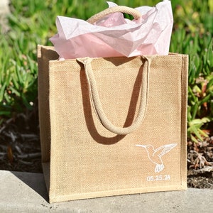 May include: A beige burlap tote bag with white handles and a white hummingbird design. The bag has the date 05.25.24 printed on the bottom.