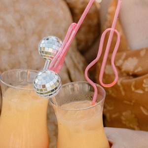 May include: Two orange and pink cocktails with disco ball straws. The cocktails are in tall, clear glasses and are being held by two people.