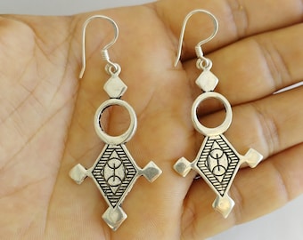 May include: A pair of silver-toned drop earrings featuring a geometric design with a circular top connected to a diamond-shaped base. The base includes engraved horizontal lines and a central oval motif with a vertical line, finished with hook wires.