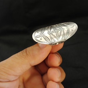 May include: A silver-colored metal ring featuring an elongated, oval-shaped face. The surface of the ring is decorated with engraved geometric patterns, including cross-hatched lines and star-like motifs arranged in a repeating triangular grid.