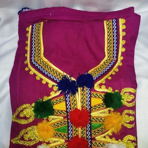May include: A pink fabric garment with gold embroidery and colorful pom-poms. The embroidery features a geometric pattern with zig-zag lines and a central design.