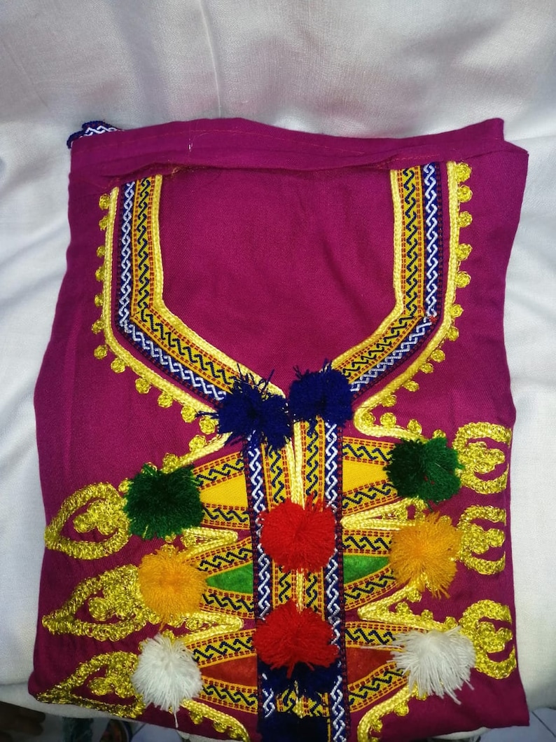 May include: A pink fabric garment with gold embroidery and colorful pom-poms. The embroidery features a geometric pattern with zig-zag lines and a central design.