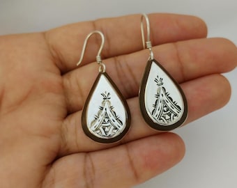 May include: A pair of silver teardrop earrings with a detailed tribal design. The earrings have a silver interior with a dark brown border and a hook closure. The earrings are displayed against a neutral background.