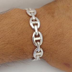 May include: A silver chain link bracelet. The bracelet features a series of connected oval links, each with a central bar. The links are smooth and shiny, reflecting light. The bracelet is worn on a wrist, showcasing its design and craftsmanship.