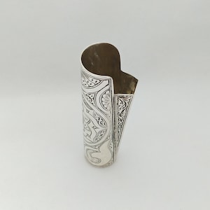 May include: Silver cigarette lighter with an intricate engraved design. The lighter is cylindrical and has a hinged lid.