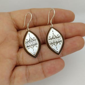 May include: A pair of silver earrings in a teardrop shape. The earrings feature a silver interior with a black geometric design and a dark brown border. The earrings are shown on a hand.