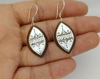 May include: A pair of silver earrings with a teardrop shape. The earrings have a silver interior with a black geometric design and a dark brown border. The earrings are displayed on a hand.