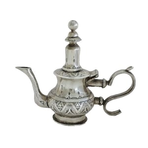 Könnte beinhalten: Eine silberne Teekanne mit einem verzierten Design und einem Ausguss an der Seite. Die Teekanne hat einen runden Korpus und einen hohen, schlanken Deckel mit einem Knauf oben.